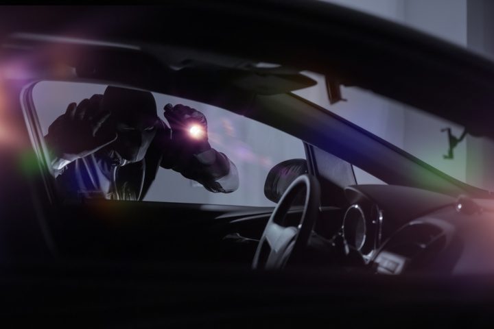 A person dressed in black looking through the window of a car with a torch
